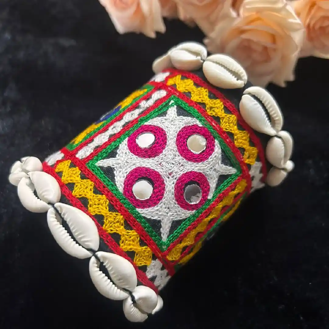Close-up of a handcrafted multicolored fabric cuff featuring intricate mirror work embroidery and a border of white cowrie shells on a black background