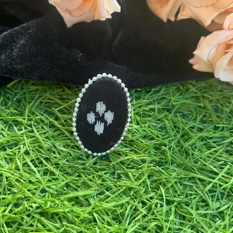 A handcrafted black fabric oval statement ring with white floral embroidery and a silver bead border, displayed on green grass with black velvet and peach roses in the background.