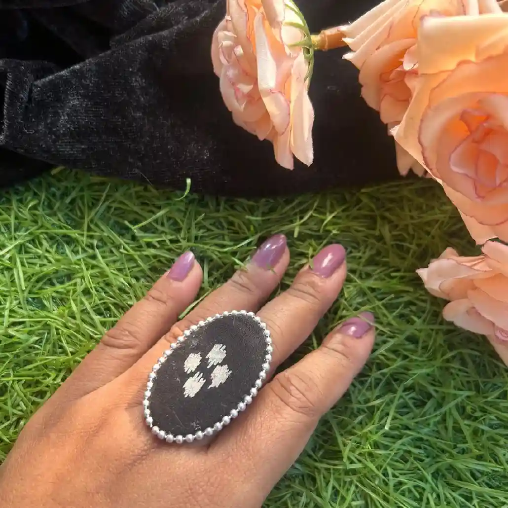 A person wearing a large handcrafted oval statement ring made of black fabric with white embroidery and a silver bead border.