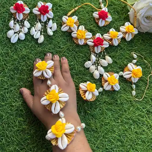 A complete handcrafted jewelry set for Haldi or Mehendi ceremonies featuring white cowrie shells with yellow and red fabric roses, including a necklace, earrings, maangtika, and a matching haathphool worn on a hand, displayed on lush green grass.
