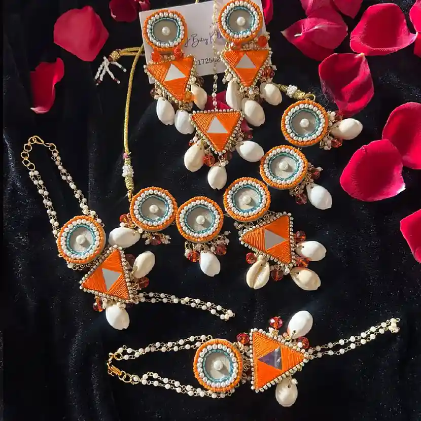 A flatlay of a handcrafted jewelry set featuring orange thread-work triangles, teal circular accents, mirror work, and white cowrie shells on black velvet with red rose petals.