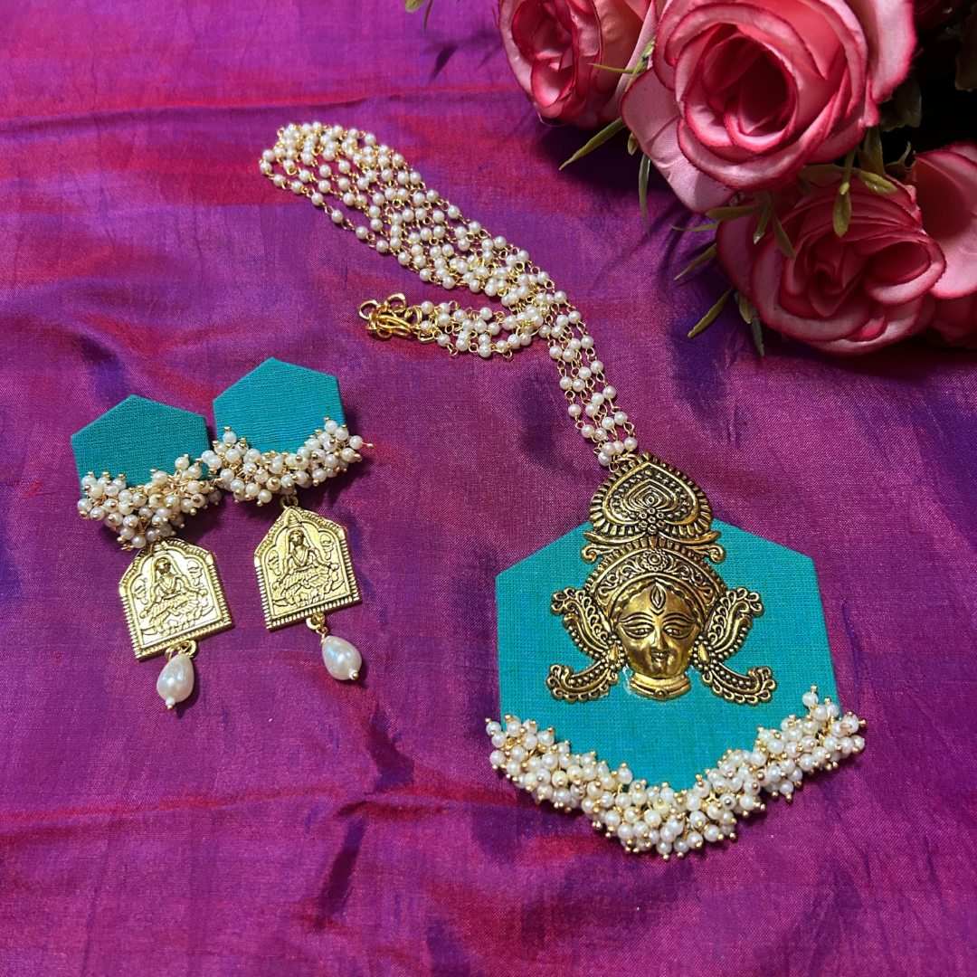 Handcrafted Durga pendant jewelry set by Itsy Bitsy Kolkata featuring a turquoise fabric base, golden Goddess Durga face motif, multi-strand pearl chain, and matching drop earrings