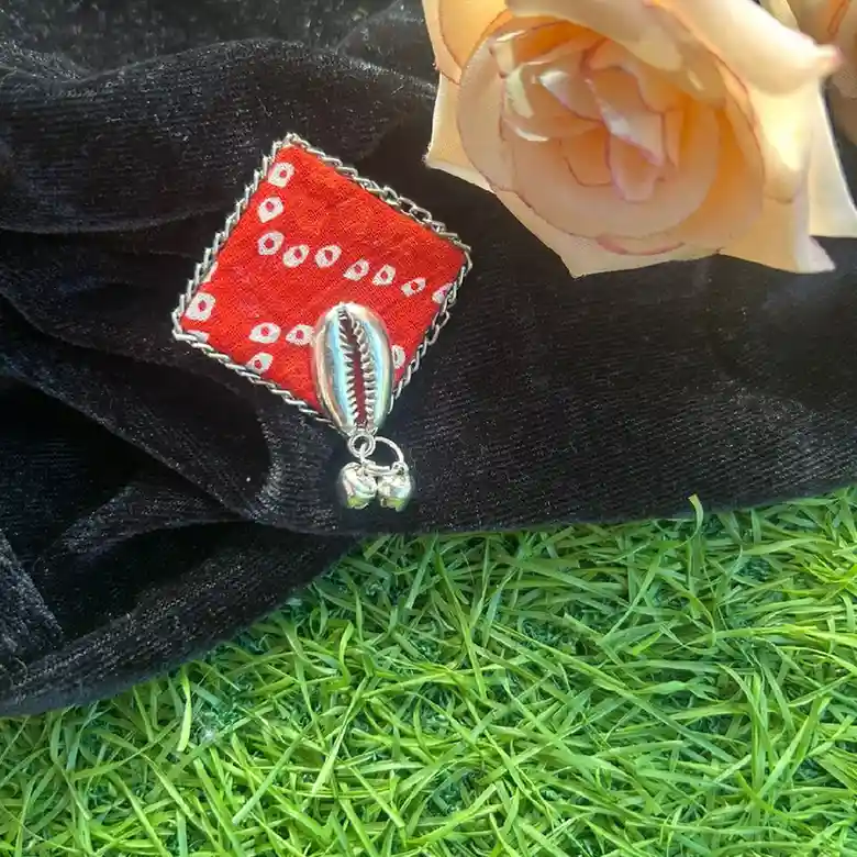 A handcrafted square statement ring made of red Bandhani fabric featuring a silver cowrie shell and hanging ghungroo beads, displayed on black velvet with a peach rose