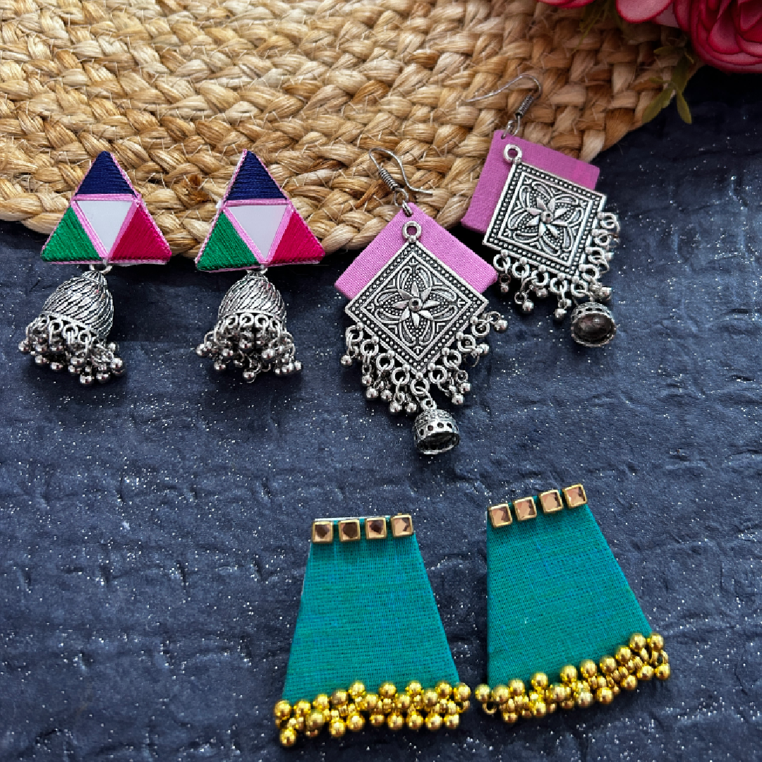A three-pair combo pack of handmade festive fabric earrings featuring colorful triangle mirror jhumkas, pink square oxidized silver mandala danglers, and teal trapeze studs with golden bead fringe.